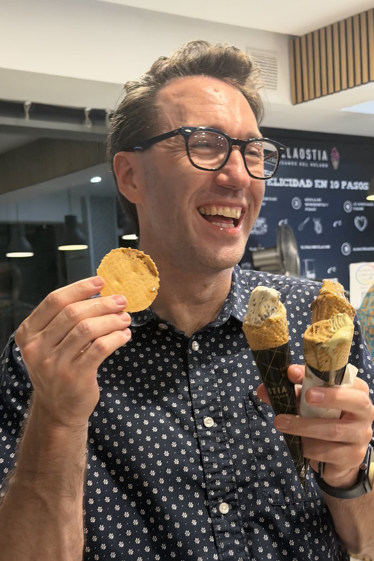 JB laughing, holding two gelato cones and a spare wafer, in a gelateria in Spain